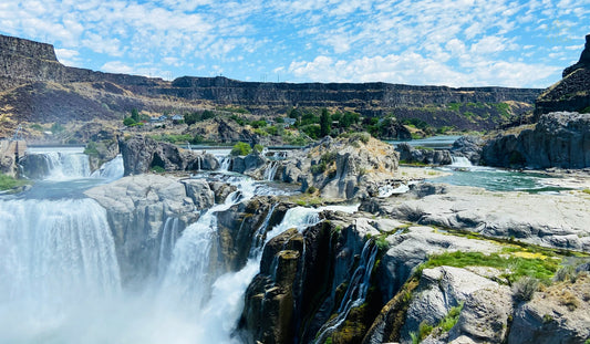 Wide multi-channel waterfall flowing over a basalt canyon at Shoshone Falls, Idaho, with rising mist and blue sky.